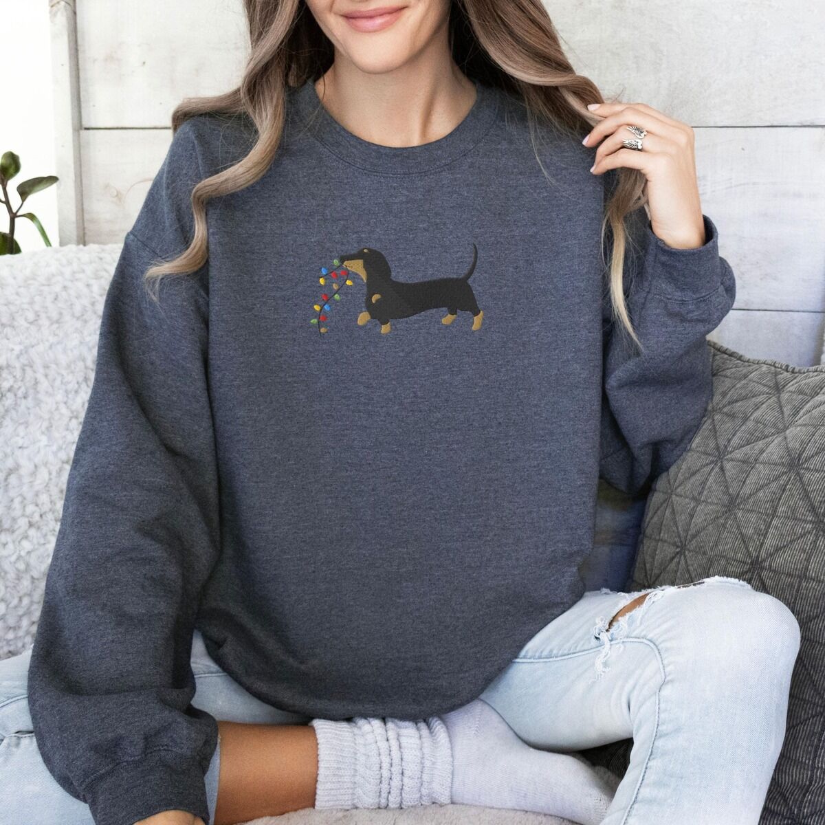 Dachshund Christmas Lights Sweatshirt – Dark Heather Woman sitting on a sofa in a Dark Heather crewneck sweatshirt, embroidered with a black and tan dachshund trotting with a string of colored Christmas lights in its mouth
