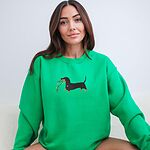Smiling woman sitting in an Irish Green crewneck sweatshirt, embroidered with a black and tan dachshund trotting with a string of colored Christmas lights in its mouth