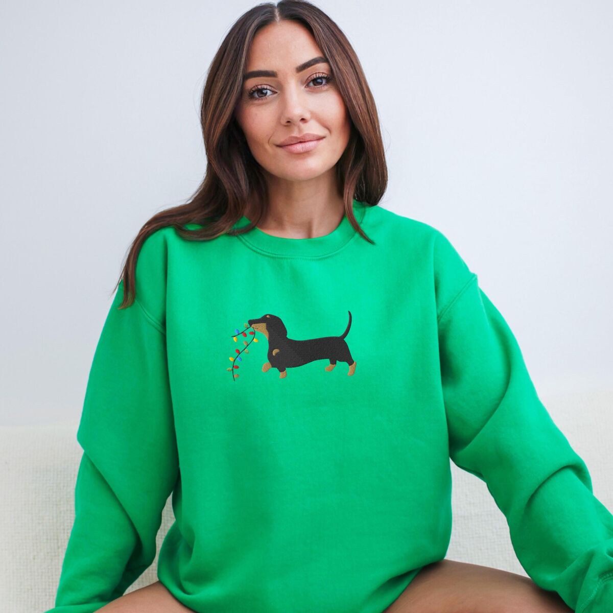 Dachshund Christmas Lights Sweatshirt - Irish Green Smiling woman sitting in an Irish Green crewneck sweatshirt, embroidered with a black and tan dachshund trotting with a string of colored Christmas lights in its mouth