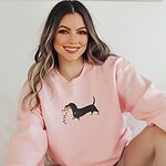 Smiling woman sitting in a Light Pink crewneck sweatshirt, embroidered with a black and tan dachshund trotting with a string of colored Christmas lights in its mouth