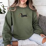 Woman sitting on a sofa in a Military Green crewneck sweatshirt, embroidered with a black and tan dachshund trotting with a string of colored Christmas lights in its mouth