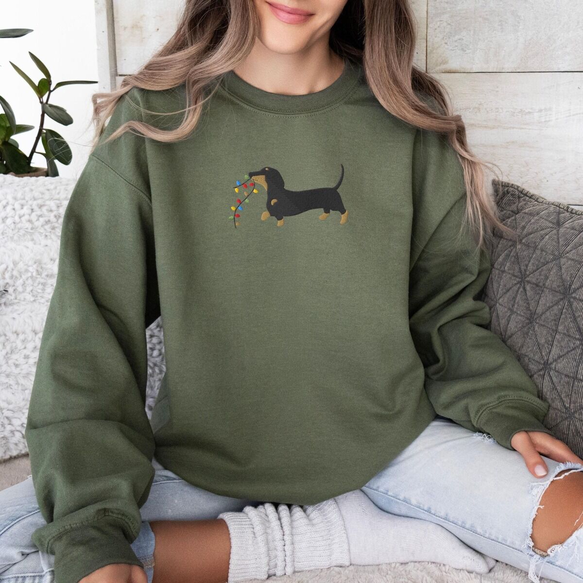 Dachshund Christmas Lights Sweatshirt – Military Green Woman sitting on a sofa in a Military Green crewneck sweatshirt, embroidered with a black and tan dachshund trotting with a string of colored Christmas lights in its mouth