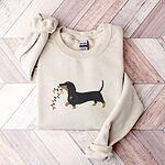 Sand crewneck sweatshirt laid flat on a wood floor, embroidered with a black and tan dachshund trotting with a string of colored Christmas lights in its mouth