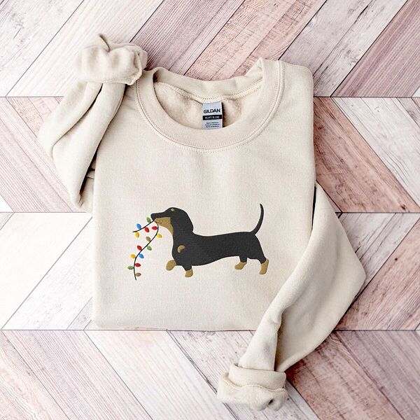 Sand crewneck sweatshirt laid flat on a wood floor, embroidered with a black and tan dachshund trotting with a string of colored Christmas lights in its mouth
