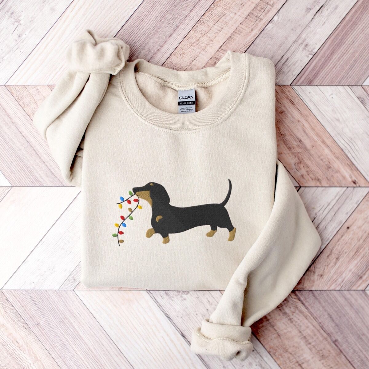 Dachshund Christmas Lights Sweatshirt – Sand Sand crewneck sweatshirt laid flat on a wood floor, embroidered with a black and tan dachshund trotting with a string of colored Christmas lights in its mouth