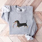 Sport Grey crewneck sweatshirt laid flat on a wood floor, embroidered with a black and tan dachshund trotting with a string of colored Christmas lights in its mouth