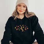 Woman in a beanie wearing a Black crewneck sweatshirt embroidered with a red dachshund sitting up on its hind legs among orange, red and gold autumn leaves across the chest