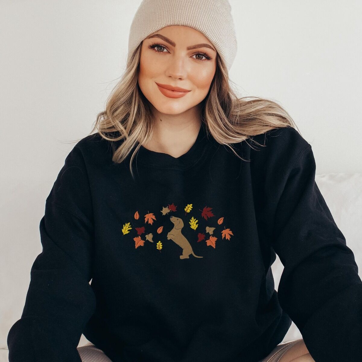 Dachshund Fall Sweatshirt – Black Woman in a beanie wearing a Black crewneck sweatshirt embroidered with a red dachshund sitting up on its hind legs among orange, red and gold autumn leaves across the chest