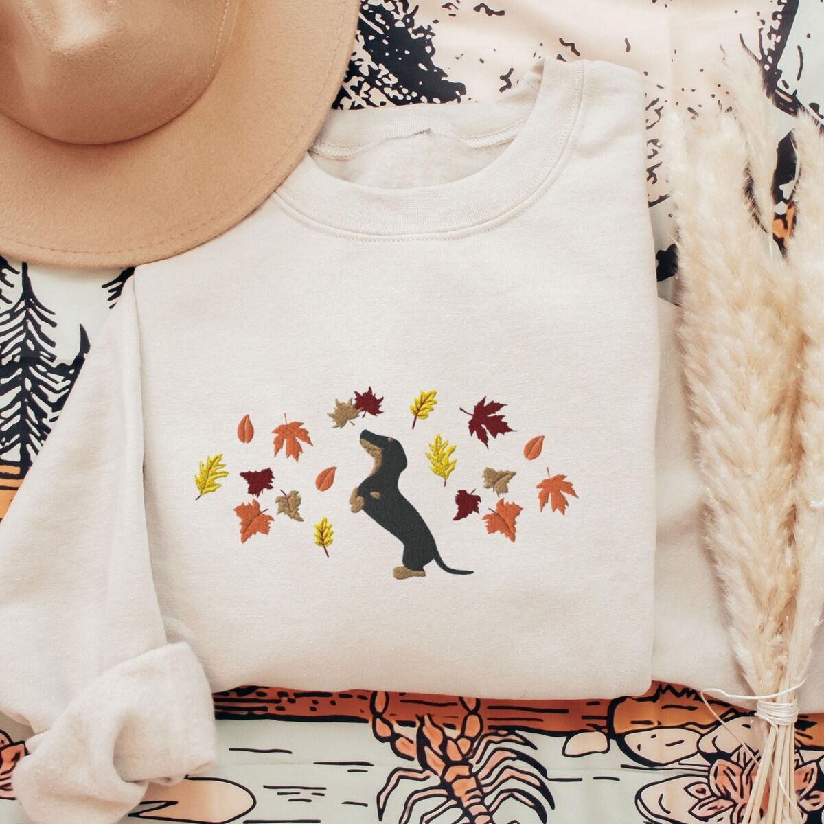 Dachshund Fall Sweatshirt - Sand with a hat Sand crewneck sweatshirt laid flat with a hat and pampas grass, embroidered with a black and tan dachshund sitting up on its hind legs among orange, red and gold autumn leaves across the chest
