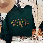 Forest Green crewneck sweatshirt laid flat with a hat and pampas grass, embroidered with a red dachshund sitting up on its hind legs among orange, red and gold autumn leaves across the chest