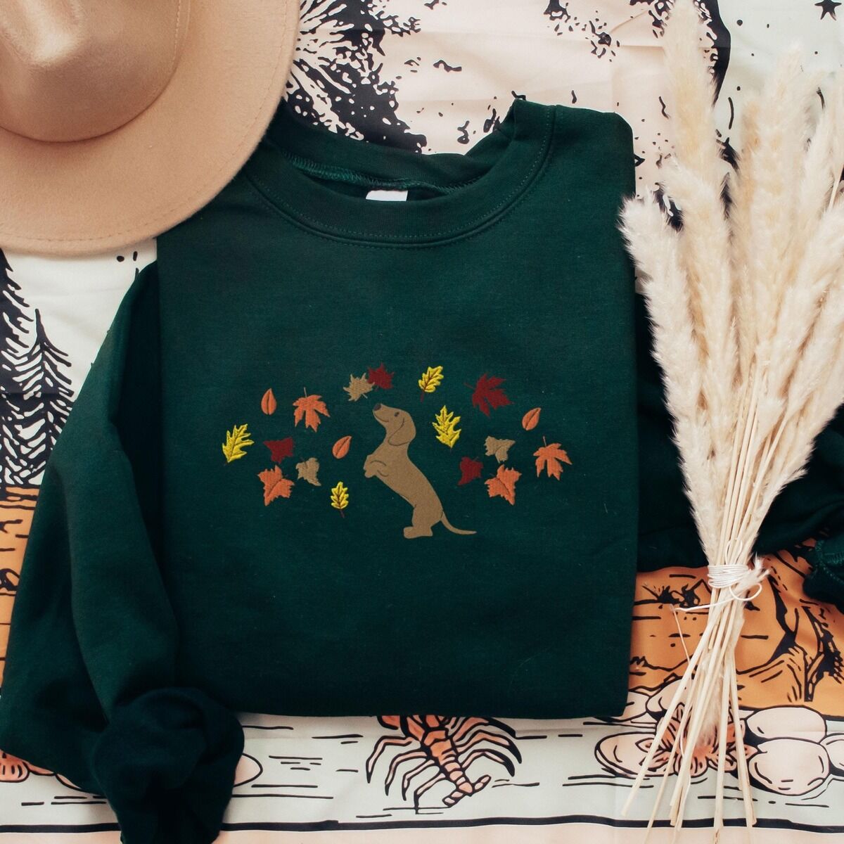 Dachshund Fall Sweatshirt – Forest Green Forest Green crewneck sweatshirt laid flat with a hat and pampas grass, embroidered with a red dachshund sitting up on its hind legs among orange, red and gold autumn leaves across the chest