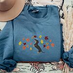 Indigo Blue crewneck sweatshirt laid flat with a hat and pampas grass, embroidered with a black and tan dachshund sitting up on its hind legs among orange, red and gold autumn leaves across the chest