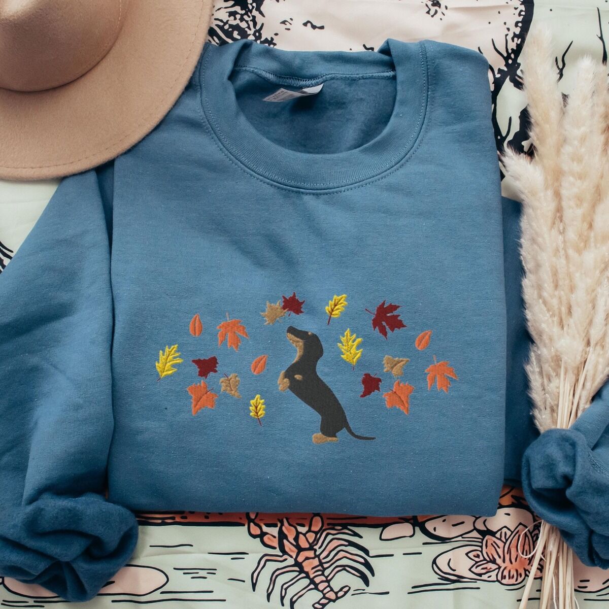 Dachshund Fall Sweatshirt – Indigo Blue Indigo Blue crewneck sweatshirt laid flat with a hat and pampas grass, embroidered with a black and tan dachshund sitting up on its hind legs among orange, red and gold autumn leaves across the chest