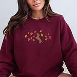 Woman in a Maroon crewneck sweatshirt embroidered with a red dachshund sitting up on its hind legs among orange, red and gold autumn leaves across the chest