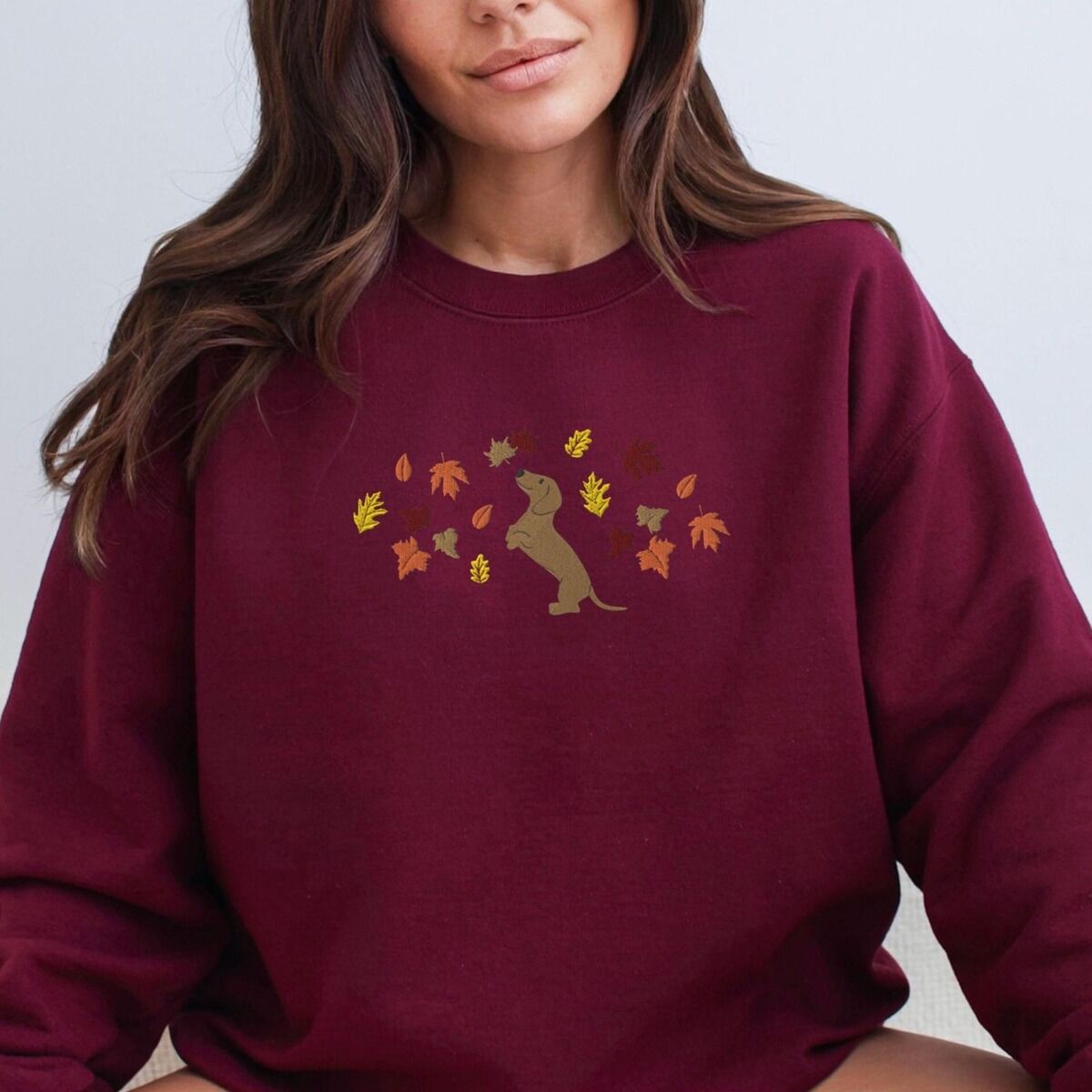 Dachshund Fall Sweatshirt – Maroon Woman in a Maroon crewneck sweatshirt embroidered with a red dachshund sitting up on its hind legs among orange, red and gold autumn leaves across the chest