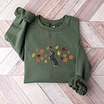 Military Green crewneck sweatshirt laid flat on herringbone wood, embroidered with a black and tan dachshund sitting up on its hind legs among orange, red and gold autumn leaves across the chest