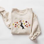 Sand crewneck sweatshirt laid flat on white, embroidered with a black and tan dachshund sitting up on its hind legs among orange, red and gold autumn leaves across the chest