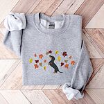 Sport Grey crewneck sweatshirt laid flat on herringbone wood, embroidered with a black and tan dachshund sitting up on its hind legs among orange, red and gold autumn leaves across the chest