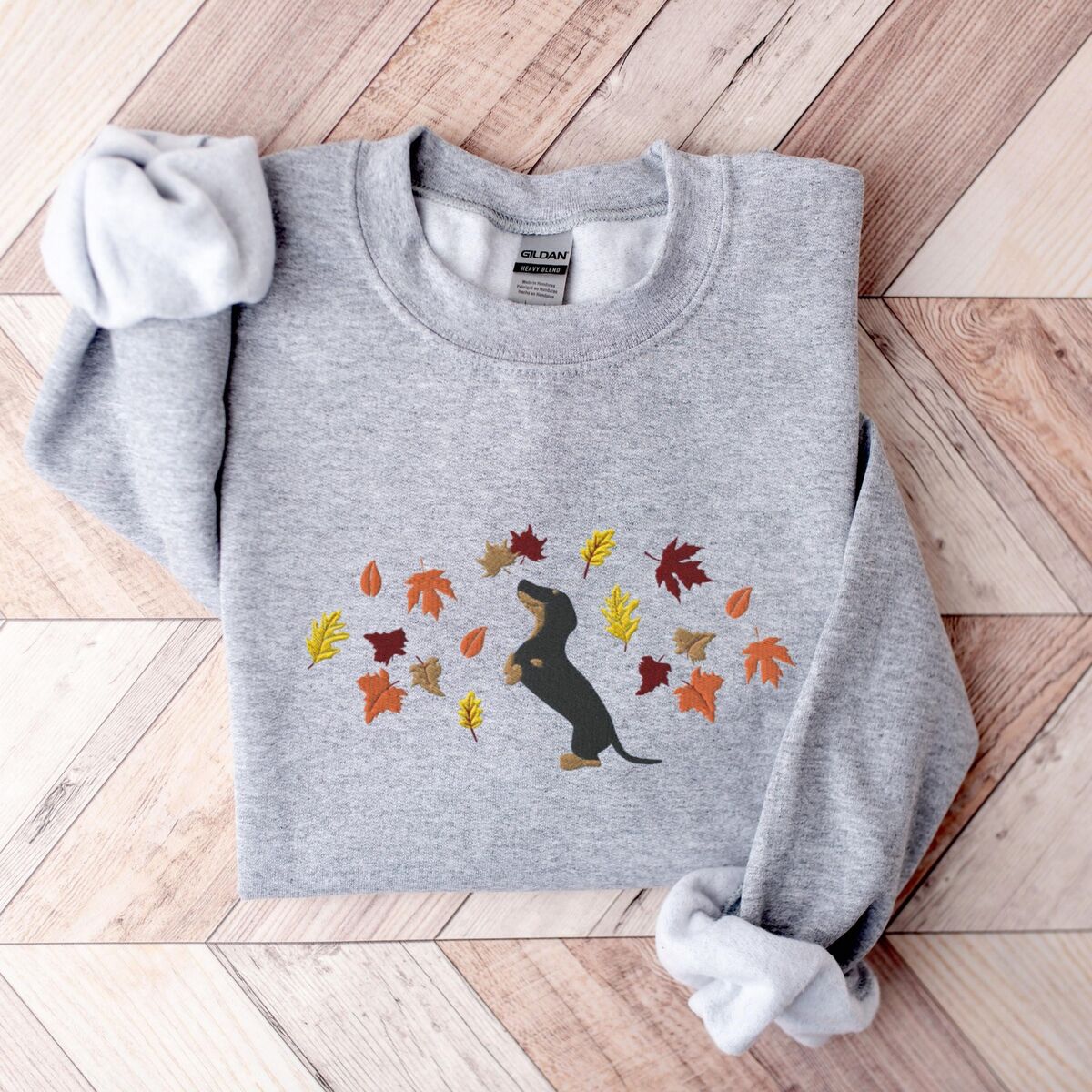 Dachshund Fall Sweatshirt - Sport Grey Sport Grey crewneck sweatshirt laid flat on herringbone wood, embroidered with a black and tan dachshund sitting up on its hind legs among orange, red and gold autumn leaves across the chest