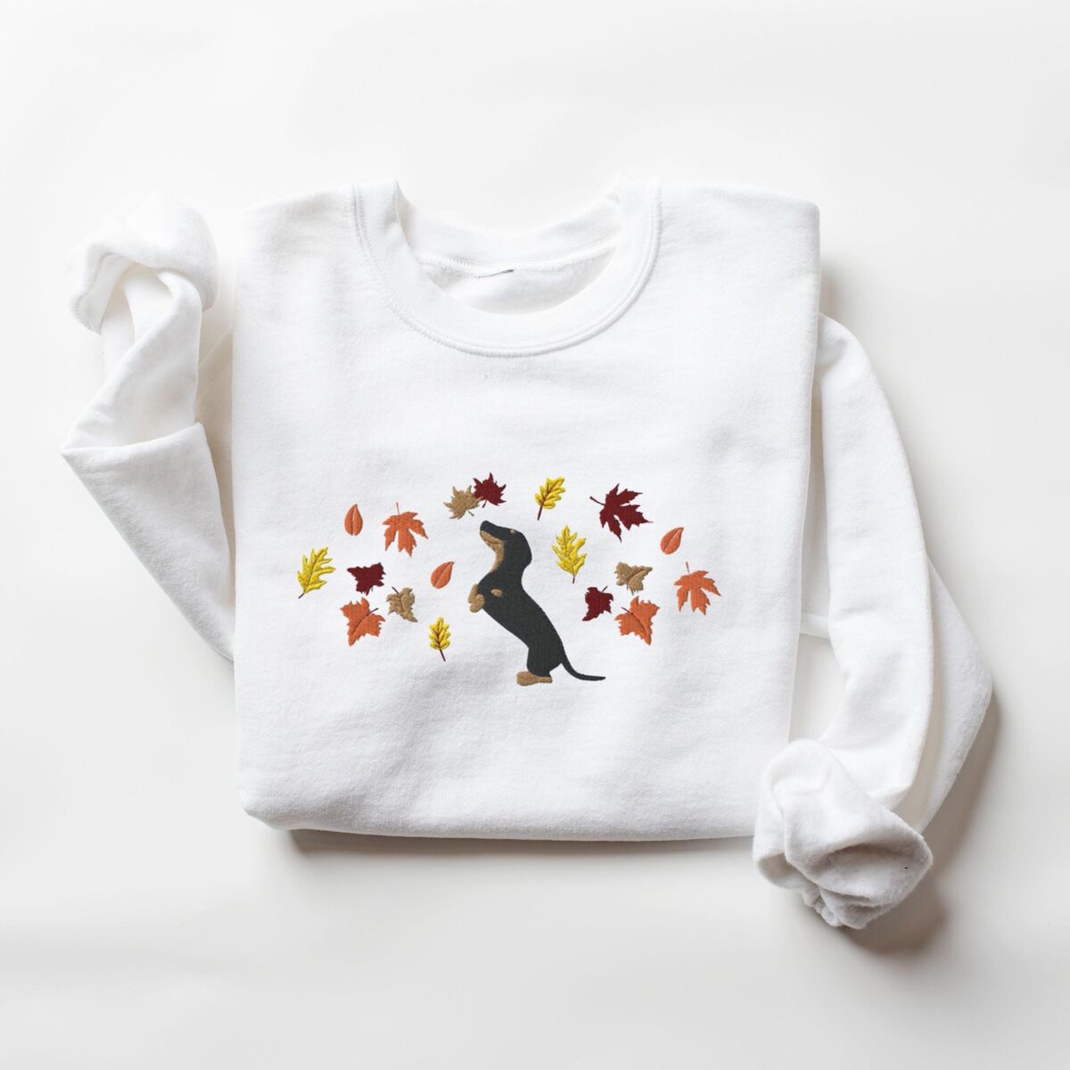 Dachshund Fall Sweatshirt - White White crewneck sweatshirt laid flat on white, embroidered with a black and tan dachshund sitting up on its hind legs among orange, red and gold autumn leaves across the chest