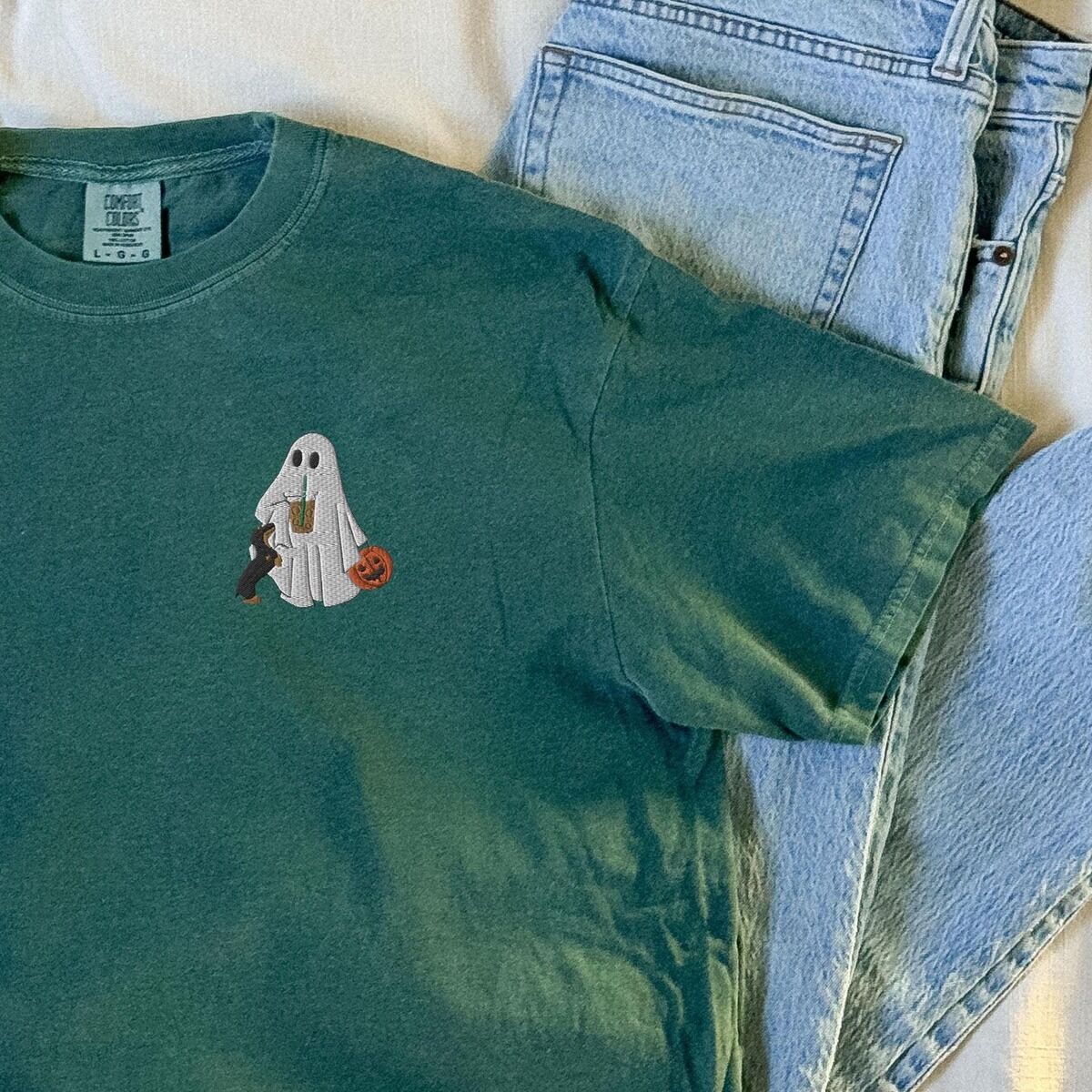 Dachshund Ghost Coffee Shirt – Blue Spruce Blue Spruce T-shirt laid on a bed beside folded jeans, embroidered on the left chest with a ghost sipping iced coffee, a black and tan dachshund and a jack-o'-lantern pail