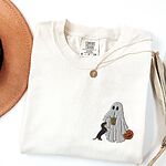 Ivory T-shirt folded flat with a necklace, a brown hat and dried palm leaves, embroidered with a ghost sipping iced coffee, a black and tan dachshund at her side and a jack-o'-lantern pail on the left chest