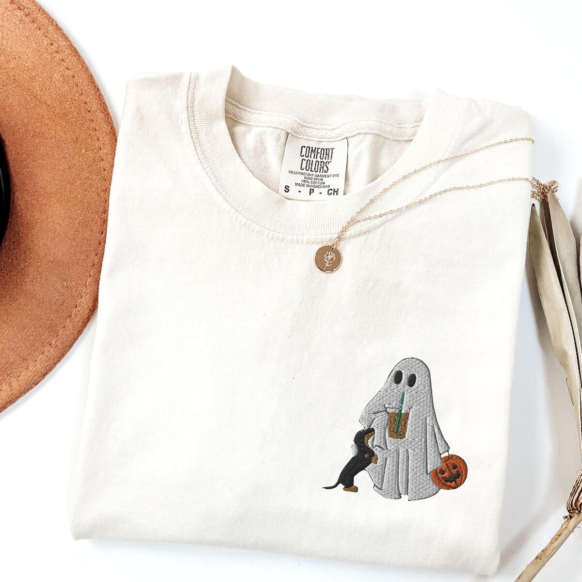 Dachshund Ghost Coffee Shirt – Ivory Ivory T-shirt folded flat with a necklace, a brown hat and dried palm leaves, embroidered with a ghost sipping iced coffee, a black and tan dachshund at her side and a jack-o'-lantern pail on the left chest