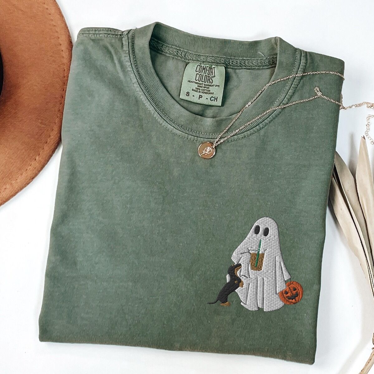 Dachshund Ghost Coffee Shirt – Moss Moss T-shirt folded flat with a necklace, a brown hat and dried palm leaves, embroidered with a ghost sipping iced coffee, a black and tan dachshund at her side and a jack-o'-lantern pail on the left chest