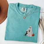 Seafoam T-shirt folded flat with a necklace, a brown hat and dried palm leaves, embroidered with a ghost sipping iced coffee, a black and tan dachshund at her side and a jack-o'-lantern pail on the left chest