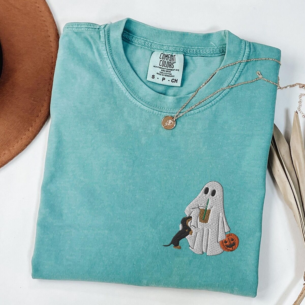 Dachshund Ghost Coffee Shirt – Seafoam Seafoam T-shirt folded flat with a necklace, a brown hat and dried palm leaves, embroidered with a ghost sipping iced coffee, a black and tan dachshund at her side and a jack-o'-lantern pail on the left chest