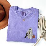 Violet T-shirt folded flat with a necklace, a brown hat and dried palm leaves, embroidered with a ghost sipping iced coffee, a black and tan dachshund at her side and a jack-o'-lantern pail on the left chest