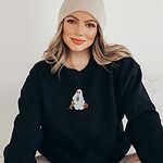 Woman sitting in a Black crewneck sweatshirt embroidered with a ghost sipping iced coffee, a red dachshund standing up at her side and a jack-o'-lantern pail