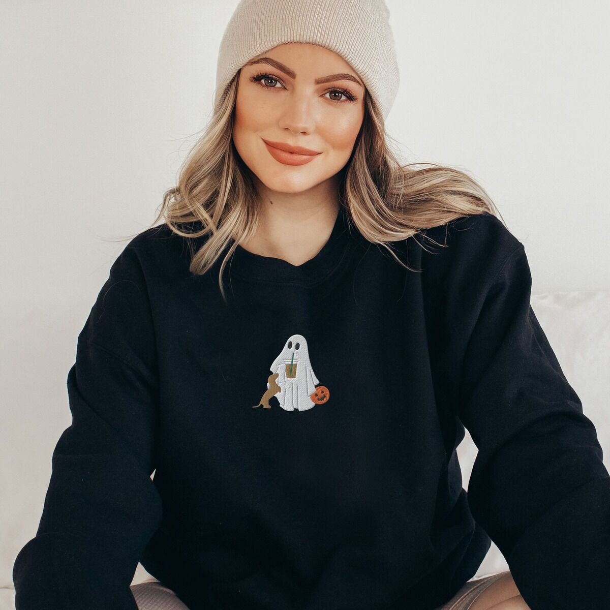 Dachshund Ghost Coffee Sweatshirt – Black Woman sitting in a Black crewneck sweatshirt embroidered with a ghost sipping iced coffee, a red dachshund standing up at her side and a jack-o'-lantern pail
