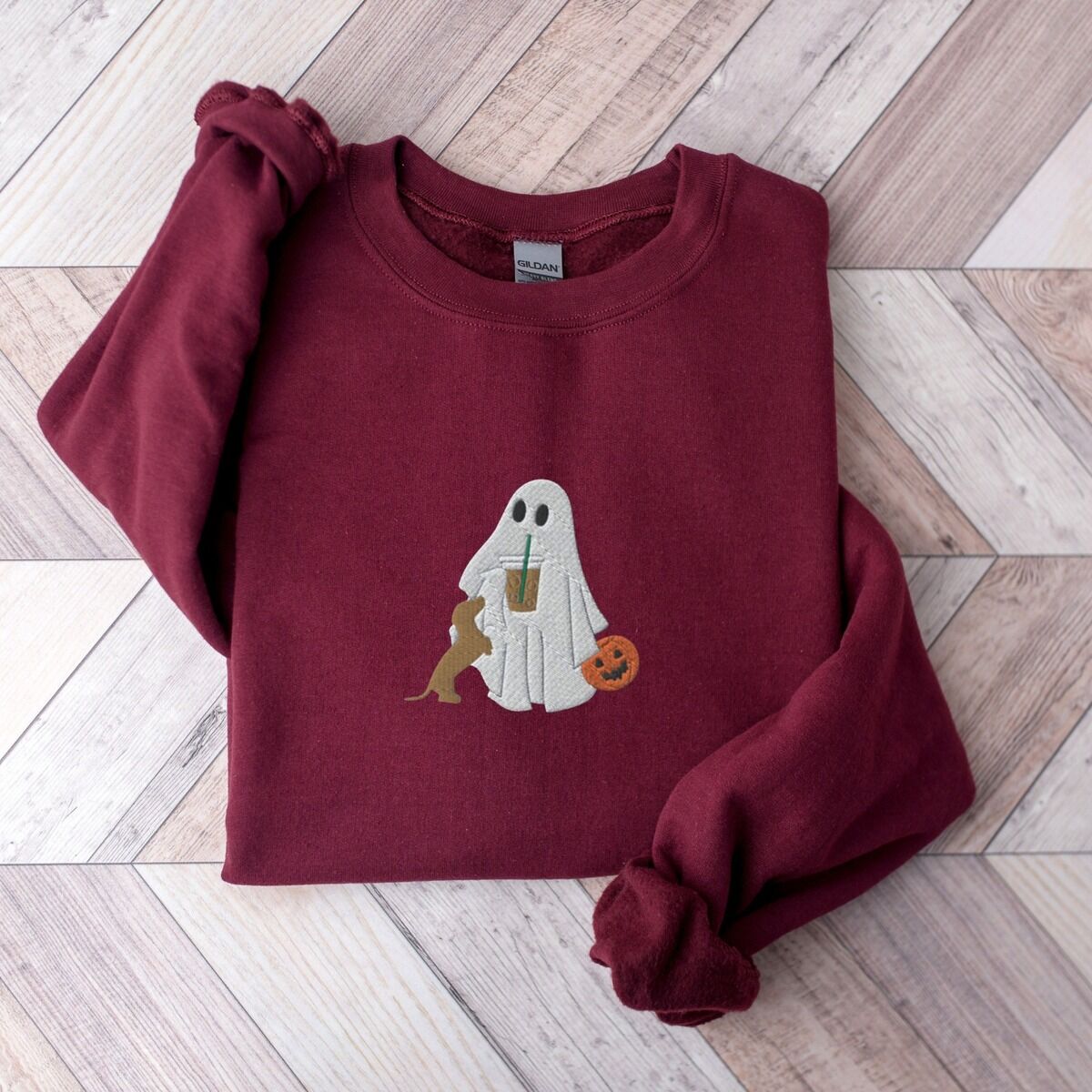 Dachshund Ghost Coffee Sweatshirt - red doxie on Maroon Maroon crewneck sweatshirt on a wood floor, embroidered with a ghost sipping iced coffee, a red dachshund standing up at her side and a jack-o'-lantern pail