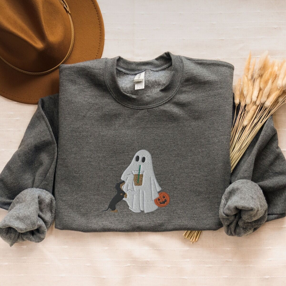 Dachshund Ghost Coffee Sweatshirt – Dark Heather Dark Heather crewneck sweatshirt laid flat with a brown hat and wheat stems, embroidered with a ghost sipping iced coffee, a black and tan dachshund standing up at her side and a jack-o'-lantern pail