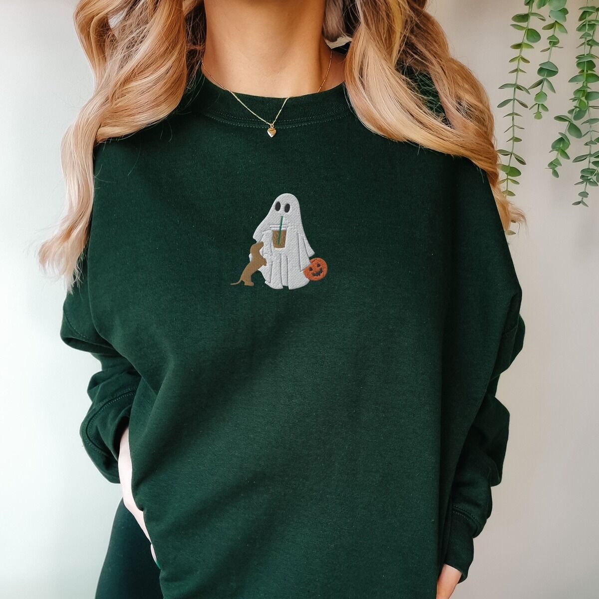 Dachshund Ghost Coffee Sweatshirt – Forest Green Woman in a Forest Green crewneck sweatshirt embroidered with a ghost sipping iced coffee, a red dachshund standing up at her side and a jack-o'-lantern pail