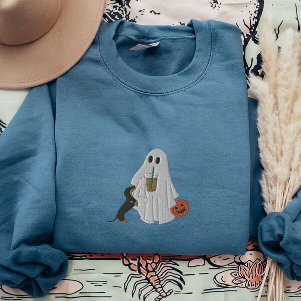 Indigo Blue crewneck sweatshirt laid flat with a brown hat and dried pampas grass, embroidered with a ghost sipping iced coffee, a black and tan dachshund standing up at her side and a jack-o'-lantern pail