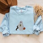 Light Blue crewneck sweatshirt laid flat with a brown hat and wheat stems, embroidered with a ghost sipping iced coffee, a black and tan dachshund standing up at her side and a jack-o'-lantern pail