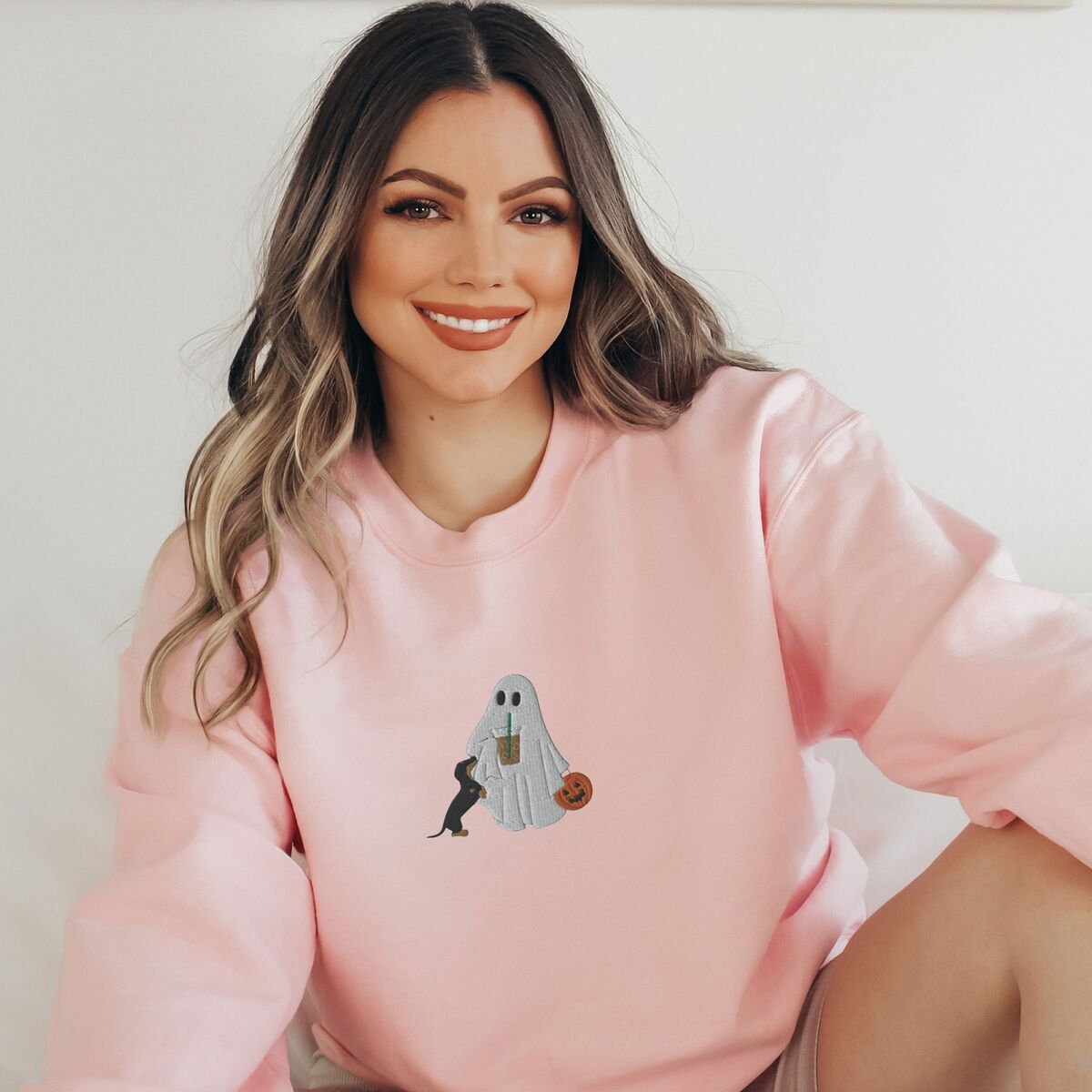 Dachshund Ghost Coffee Sweatshirt - Light Pink Smiling woman in a Light Pink crewneck sweatshirt embroidered with a ghost sipping iced coffee, a black and tan dachshund at her side and a jack-o'-lantern pail