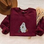 Maroon crewneck sweatshirt laid flat with a brown hat and wheat stems, embroidered with a ghost sipping iced coffee, a black and tan dachshund standing up at her side and a jack-o'-lantern pail