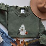 Military Green crewneck sweatshirt laid flat beside jeans, a plaid scarf and autumn leaves, embroidered with a ghost sipping iced coffee, a black and tan dachshund and a jack-o'-lantern pail