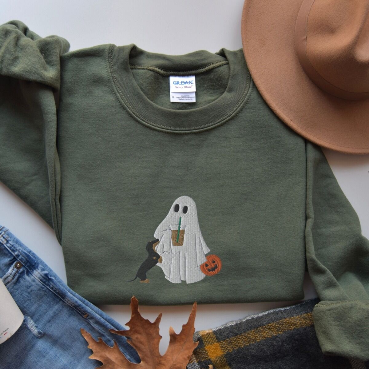 Dachshund Ghost Coffee Sweatshirt – Military Green Military Green crewneck sweatshirt laid flat beside jeans, a plaid scarf and autumn leaves, embroidered with a ghost sipping iced coffee, a black and tan dachshund and a jack-o'-lantern pail