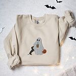 Sand crewneck sweatshirt laid flat among paper bats and fairy lights, embroidered with a ghost sipping iced coffee, a black and tan dachshund standing up at her side and a jack-o'-lantern pail
