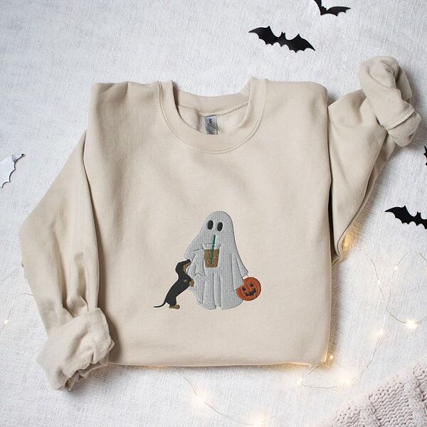 Sand crewneck sweatshirt laid flat among paper bats and fairy lights, embroidered with a ghost sipping iced coffee, a black and tan dachshund standing up at her side and a jack-o'-lantern pail