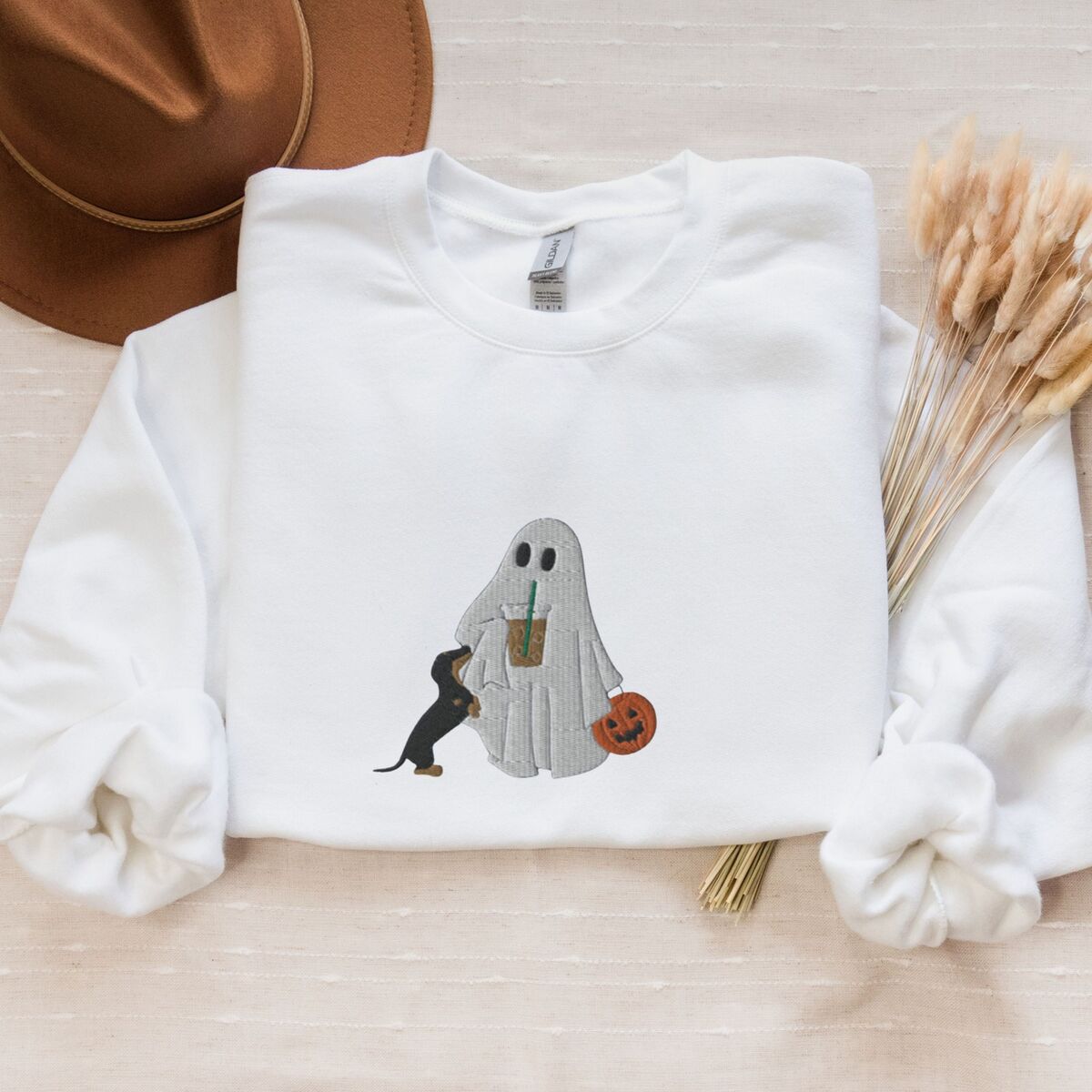 Dachshund Ghost Coffee Sweatshirt - White White crewneck sweatshirt laid flat with a brown hat and wheat stems, embroidered with a ghost sipping iced coffee, a black and tan dachshund standing up at her side and a jack-o'-lantern pail