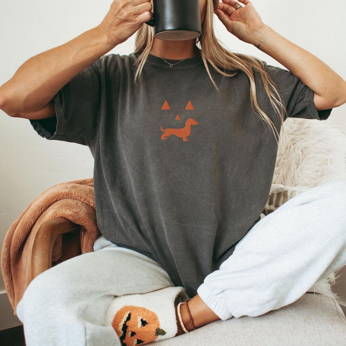 Dachshund Jack-o'-Lantern Shirt - worn Woman sipping from a mug in a dark grey T-shirt embroidered with an orange dachshund jack-o'-lantern face, a pumpkin pillow at her feet
