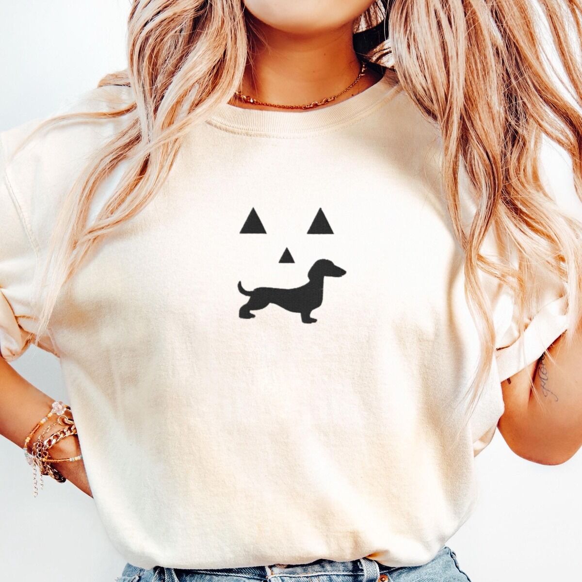 Dachshund Jack-o'-Lantern Shirt – Ivory Woman in an Ivory T-shirt embroidered across the chest with a black jack-o'-lantern face whose grin is a dachshund silhouette