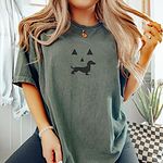 Woman in a Moss T-shirt embroidered across the chest with a black jack-o'-lantern face with a dachshund silhouette smile