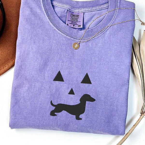 Violet T-shirt laid flat with a necklace, embroidered across the chest with a black jack-o'-lantern face: triangle eyes and nose over a dachshund silhouette grin