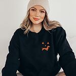 Woman in a beanie wearing a Black crewneck sweatshirt embroidered on the left chest with an orange jack-o'-lantern face whose grin is a dachshund silhouette